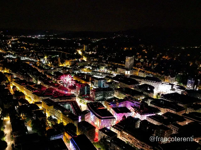 Night over the lit city, a red Ferris wheel glowing above the rooftops while whole blocks are washed in magenta, violet and red light. The station's illuminated clock tower, a glass office high rise and a floodlit church spire stand out among the amber street grid, with dark wooded hills closing the horizon. Aerial drone photograph over ST GALLEN, Switzerland from 47.421699641459384, 9.37512516975403 on 2025-08-01. Exposure 1970 · f1.8 · 9mm.