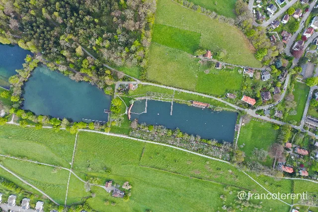Straight down over the Drei Weieren bathing ponds, their deep green water framed by timber jetties and red roofed wooden bathhouses set along the dividing dam. Woodland and a smaller upper pond fill the left, while a strip of allotment gardens, bright spring meadows crossed by footpaths and scattered houses fill the right. Aerial drone photograph over ST GALLEN, Switzerland from 47.421772232407164, 9.38035011291504 on 2024-04-27. Exposure 100 · f3.5 · 10mm.