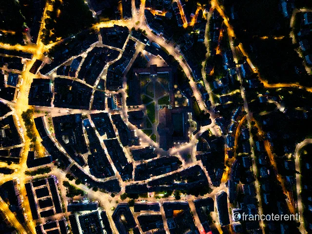 Straight down on the medieval old town at night, the abbey precinct at the centre with the cathedral's cross shaped roof and the dark lawns of the Klosterhof lit only along their paths. Warm amber streetlights trace the ring road around a tight oval of unlit rooftops and narrow lanes, with wooded slopes going black at the top right. Aerial drone photograph over ST GALLEN, Switzerland from 47.42400798458176, 9.377903938293459 on 2025-07-14. Exposure 1400 · f1.8 · 9mm.