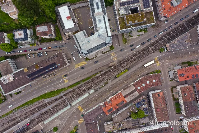 Straight down on a rail corridor cutting diagonally across the frame, tracks and a glass roofed platform running beside a wide multi lane road marked with yellow crossings. Office blocks with solar panels and green roofs sit on one side, red tiled townhouses and parked cars on the other, under flat grey light. Aerial drone photograph over ST GALLEN, Switzerland from 47.42339824340531, 9.37086582183838 on 2024-04-30. Exposure 100 · f4 · 10mm.