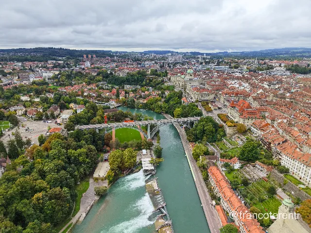 Turquoise river running between steep wooded banks, crossed by the riveted steel arch of the Kirchenfeldbrücke; below it a green sports pitch with a red gravel track, and in the foreground the Schwellenmätteli weir churning white water beside a covered riverside terrace. Beyond, the copper-domed Federal Palace stands above dense red-tiled old-town roofs, with wooded ridges and a flat grey overcast closing the horizon. Aerial drone photograph over BERN, Switzerland from 46.94346388408633, 7.44950294494629 on 2025-09-22. Exposure 220 · f1.8 · 9mm.