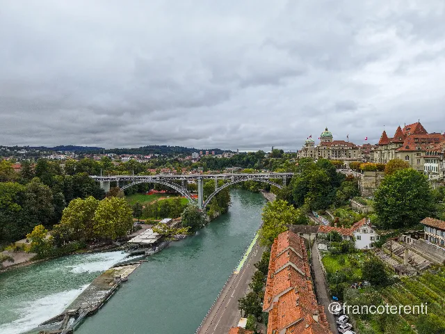 Low, heavy grey cloud over a milky turquoise river, the pale steel arch of the Kirchenfeldbrücke leaping the channel between tree-covered banks. A stepped weir spills foam past a glass-roofed riverside terrace on the left, while a long terrace of orange-tiled houses and quay road fills the right foreground, rising to the domed Federal Palace and steep-roofed old-town facades on the bluff above. Aerial drone photograph over BERN, Switzerland from 46.94920794303434, 7.45250701904297 on 2025-09-22. Exposure 170 · f1.8 · 9mm.