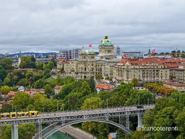 The Federal Palace and its flanking wings, green oxidised domes and Swiss flags catching the flat light of an overcast sky, seen across the treetops of a wooded riverbank; in the foreground a yellow tram and pedestrians cross the grey lattice steel arch of the Kirchenfeldbrücke, with sandstone hotel and townhouse facades, red roofs and distant wooded hills behind. Aerial drone photograph over BERN, Switzerland from 46.94674725306136, 7.444782257080079 on 2025-09-22. Exposure 160 · f2.8 · 19mm.