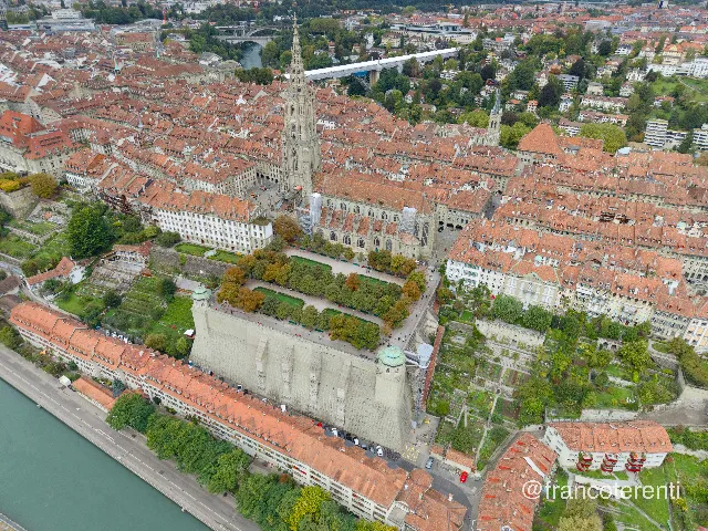 Dense mass of orange-tiled medieval roofs packed along ridge-top streets, the Gothic minster's tall openwork sandstone spire rising above them with scaffolding on its flank. Below, the Münsterplattform sits on a massive buttressed retaining wall with a domed corner pavilion, its clipped avenues of trees overlooking terraced kitchen gardens, riverside houses and the green Aare in the lower corner. Aerial drone photograph over BERN, Switzerland from 46.94856165497637, 7.458343505859376 on 2025-09-22. Exposure 100 · f1.8 · 9mm.