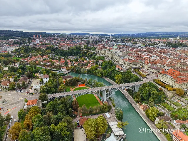 Wide sweep over a turquoise river looping past wooded banks, with the steel arch of the Kirchenfeldbrücke in the foreground above a football pitch and a smaller bridge upstream. The domed Federal Palace and the tight grid of red-roofed old-town blocks fill the right bank, terraced gardens dropping to the water, and low green hills and grey cloud stretch across the far distance. Aerial drone photograph over BERN, Switzerland from 46.94463695450445, 7.456626892089845 on 2025-09-22. Exposure 200 · f1.8 · 9mm.