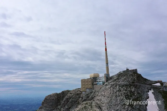 Low, level view across a bare limestone summit ridge where a beige concrete complex with a rounded rooftop pod and a tall red-and-white banded transmission mast sits on the rock, railed walkways and a small patch of leftover snow running along the crest under a flat grey overcast sky. Aerial drone photograph over SANTIS, Switzerland from 47.24369691653133, 9.352712631225588 on 2024-07-14. Exposure 100 · f7.1 · 10mm.