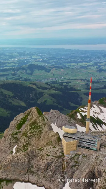 Vertical framing looking down past a beige summit building and its towering red-and-white mast clinging to a snow-streaked rock crest, with layered green ridges, scattered farms and hamlets falling away to a pale haze-covered lake and a soft blue-white cloudy sky. Aerial drone photograph over SANTIS, Switzerland from 47.24148265325081, 9.339580535888674 on 2024-07-14. Exposure 100 · f5.6 · 10mm.