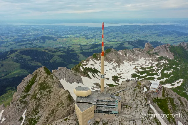 Wide elevated view over a jagged limestone crest topped by a beige complex, a rounded gold rooftop pod and a slender red-and-white mast, large snowfields filling the shaded bowl behind it while green rolling hills, pastures and villages stretch to a hazy lake on the horizon. Aerial drone photograph over SANTIS, Switzerland from 47.257271823208065, 9.344472885131838 on 2024-07-14. Exposure 100 · f5.6 · 10mm.