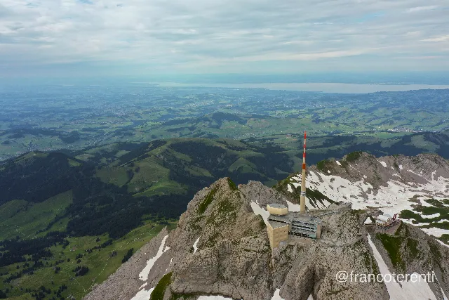 High vantage over a grey rocky summit with a beige building, terraces and a red-and-white banded mast perched on the edge, snow patches lining the ridge to the right and a vast expanse of green hills, forest and settlements fading into a pale lake and overcast sky. Aerial drone photograph over SANTIS, Switzerland from 47.25132958838516, 9.354257583618166 on 2024-07-14. Exposure 100 · f5.6 · 10mm.