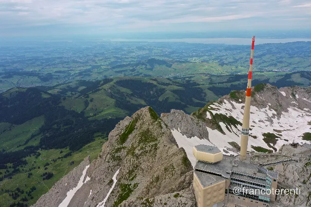 Steep rock arête with a beige summit complex and tall red-and-white mast in the lower right corner, broad melting snowfields on the shaded flank, and dark green pastures, wooded ridges and scattered farms receding to a hazy lake under thin grey cloud. Aerial drone photograph over SANTIS, Switzerland from 47.249057381208665, 9.331769943237306 on 2024-07-14. Exposure 100 · f5.6 · 10mm.